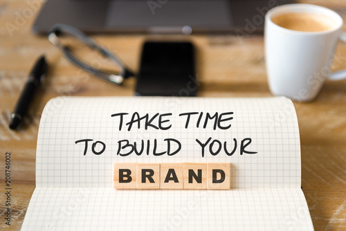 Closeup on notebook over vintage desk background, front focus on wooden blocks with letters making Take Time To Build Your Brand text