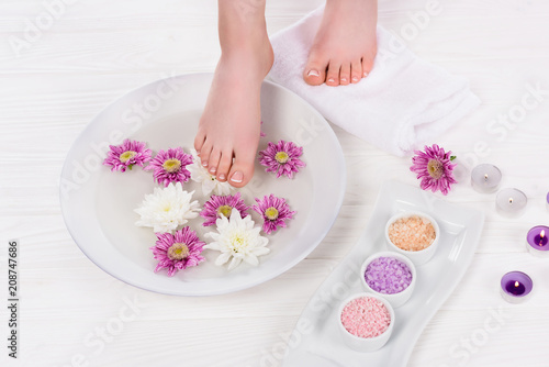 cropped shot of barefoot woman receiving bath for nails with colorful sea salt and flowers near aroma candles in beauty salon