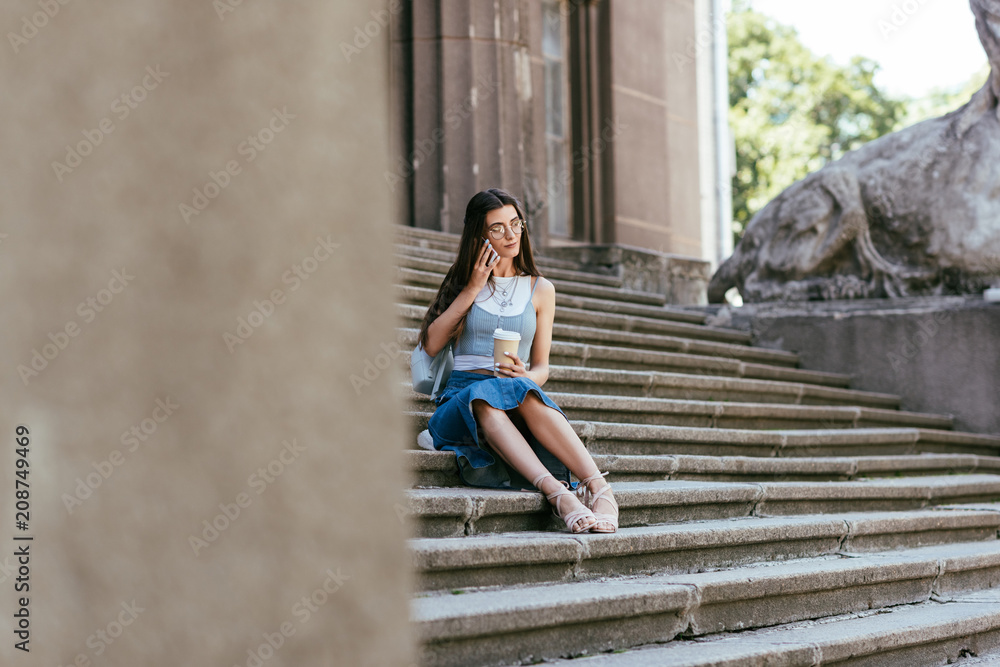 selective focus of young woman with coffee to go sitting on stairs and talking by smartphone