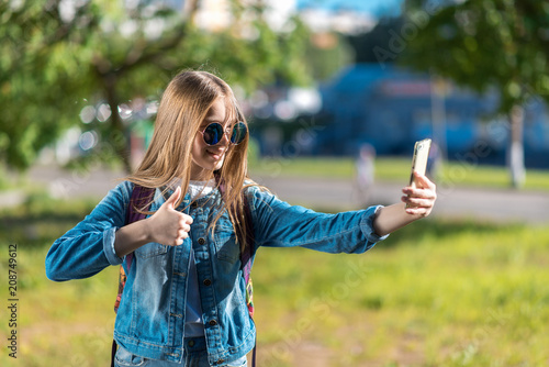 The girl is blonde. A little teenager after school. In summer in park in fresh air. In his hands holds a smartphone. A gesture of hands shows a thumbs up class. Video call with parents on smartphone.