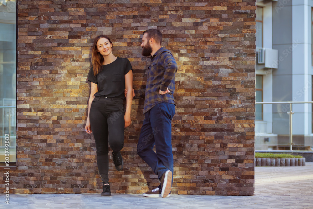 Stylish young couple is spending time together outdoors. Attractive woman and handsome bearded man are posing on brick background.