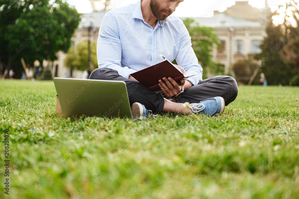 Photo of caucasian man wearing white shirt, writing down notes in notebook while sitting on grass in park with legs crossed and working on laptop