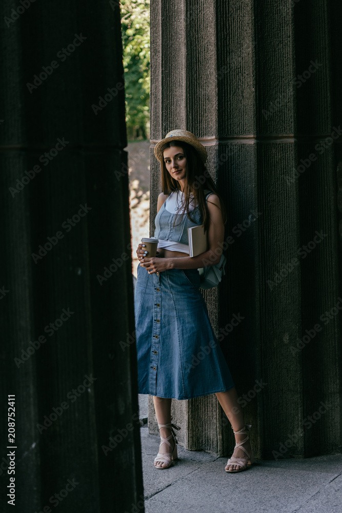 beautiful young woman with book and coffee to go standing between columns and looking at camera
