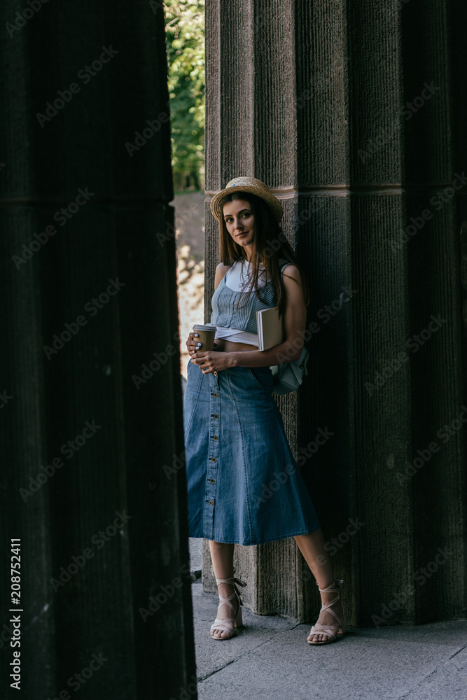 Obraz premium beautiful young woman with book and coffee to go standing between columns and looking at camera