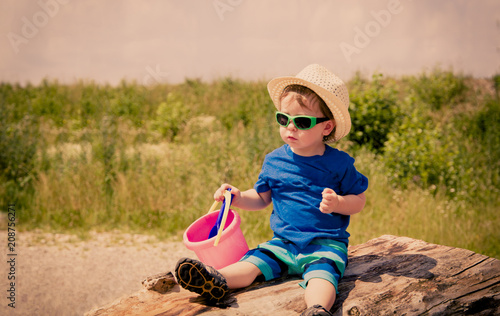 A toddler with straw hat wearing sunglasses and sand toy in hand sits on a tree trunk photo