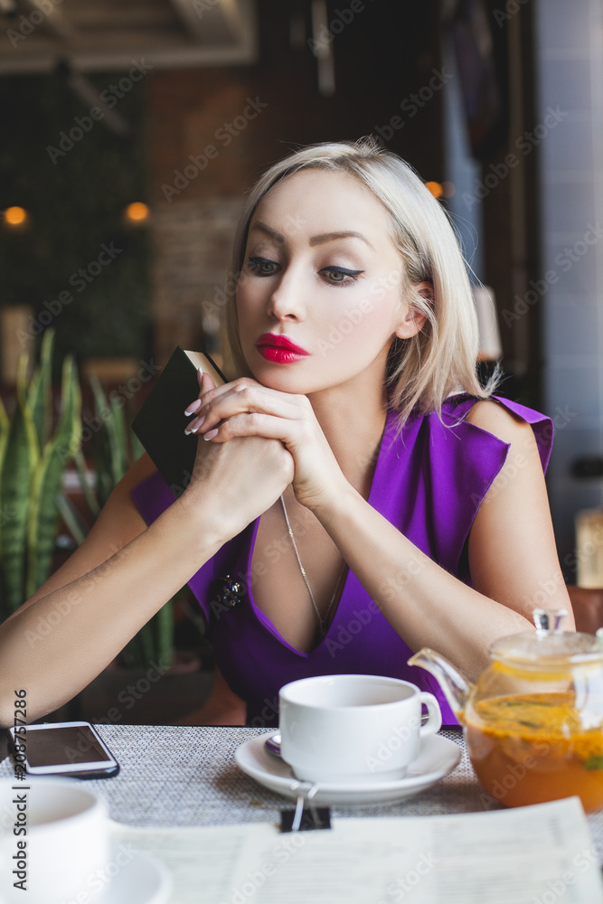 Pretty Woman sitting in Restaurant, drinking Herbal Tea, dreaming and holding Book in her Hands Pretty Woman sitting in Restaurant, drinking Herbal Tea, dreaming and holding Book in her Hands