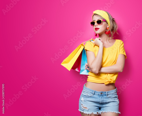 Young redhead girl in yellow t-shirt and blue jeans holding a colorful bags on pink background