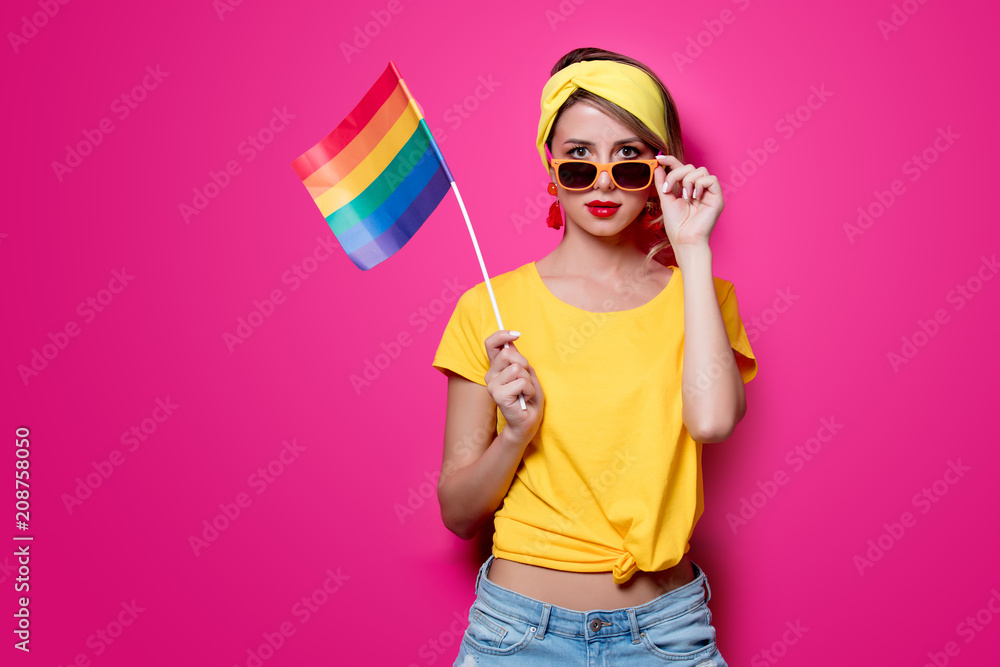 Young girl in yellow t-shirt and blue jeans holding big LGBT flag on pink background