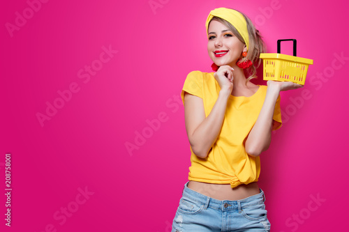 Young redhead girl in yellow t-shirt and blue jeans holding supermarket basket on pink background