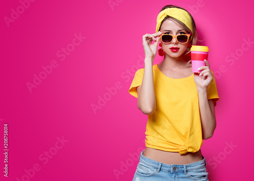 Girl in sunglasses and yellow t-shirt holding a red cup of coffee on pink background