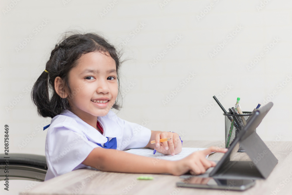 Asian little girl in student uniform doing homework and using tablet on wooden table select focus shallow depth of field