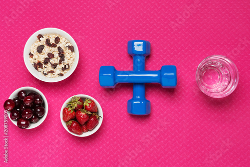 on a pink rug a plate with muesli, a pial of strawberries, a piella with cherries, dumbbells lined with a plus sign and a glass with water, a concept, a sport and a healthy lifestyle and food photo