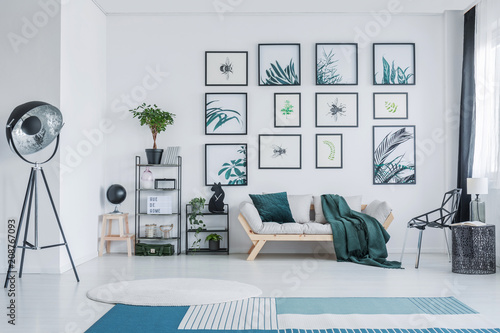 Big studio lamp standing in white living room interior with wooden sofa with green pillow and blanket, gallery with simple posters, black racks with plants and decor and two carpets on the floor