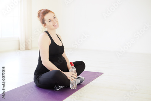 Smiling pregnant woman sitting in yoga pose with a bottle of wat
