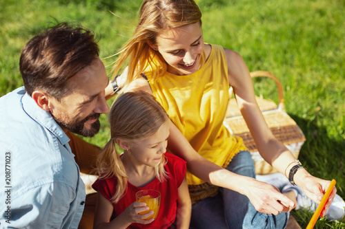 Showing pics. Cheerful blond mother holding a tablet while having a picnic with her family