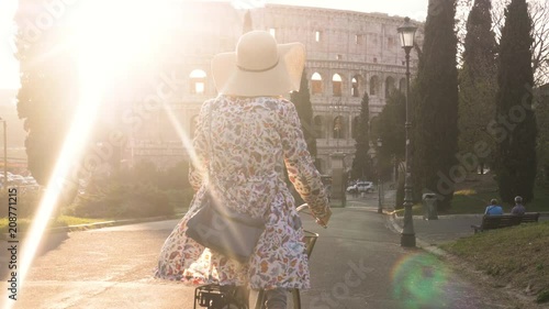 Beautiful young woman in colorful fashion dress with bike walking toward colosseum in Rome at sunset with trees happy attractive tourist girl in colle oppio photo
