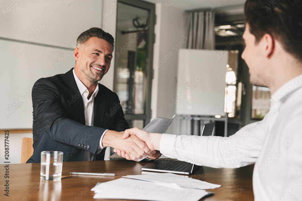 Business, career and placement concept - joyful handsome businessman 30s smiling and shaking hands with male candidate, who was recruited during interview in office - obrazy, fototapety, plakaty 