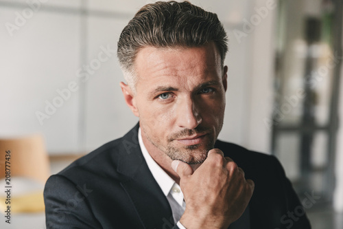 Portrait closeup of handsome businessman wearing white shirt and black suit touching chin and looking at camera, while working in office room