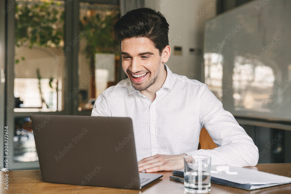 Image of handsome business man 30s wearing white shirt laughing while sitting at table in office, and looking at laptop Image of handsome business man 30s wearing white shirt laughing while sitting at table in office, and looking at laptop
