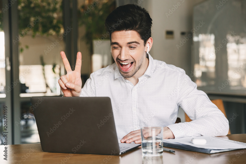 Portrait of joyful office man 30s wearing white shirt laughing and showing peace sign at laptop, during video conference or call using bluetooth earbud Portrait of joyful office man 30s wearing white shirt laughing and showing peace sign at laptop, during video conference or call using bluetooth earbud