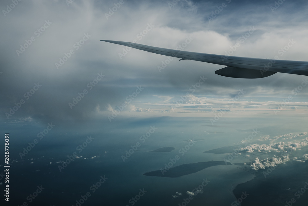 Naklejka premium Wing of airplane above island while the sun is going down with lots of clouds in the sky. Traveling concept