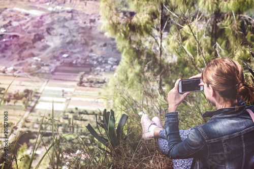 Successful woman hiker taking picture with smartphone at cliff edge on mountain top. Bali island. photo