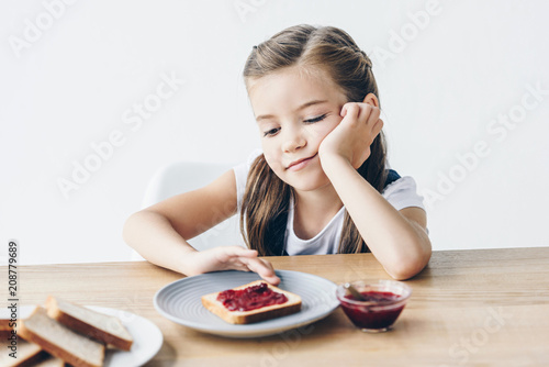 bored little schoolgirl with toasts and jam for breakfast isolated on white