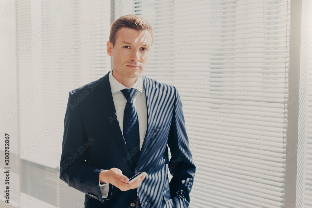 Prosperous male director sends message and multimedia files via smart phone, synchronizes phone data and shares photos for project, poses in office, wears luxury expensive suit, looks seriously Prosperous male director sends message and multimedia files via smart phone, synchronizes phone data and shares photos for project, poses in office, wears luxury expensive suit, looks seriously