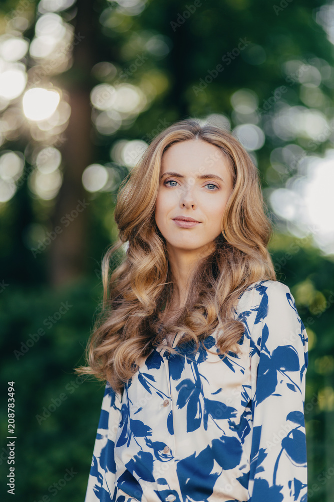 Adorable light haired young woman with charming appearance, dressed elegantly, stands outdoor against green blurred background, has walk with best friend, enjoys calm atmosphere in summer park