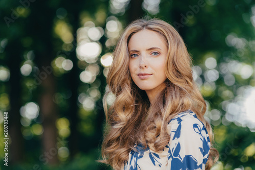 Outdoor shot of beautiful European woman has light curly hair, pure skin and blue eyes, dressed in fashionable summer dress, poses in park, enjoys good weather and sunshine. People and free time #208783448