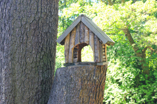 Tree House Wooden Small Handmade Birdfeeder at the Forest. Tree House and Bird Feeder Isolated on Summer Green Trees Background Close Up View. Empty Birdhouse Made from Wood Outdoors at the Park.