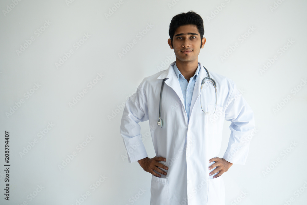 Content successful general practitioner in lab coat holding hands on hips and looking at camera. Serious confident handsome Indian male doctor working in hospital. Healthcare and medicine concept Content successful general practitioner in lab coat holding hands on hips and looking at camera. Serious confident handsome Indian male doctor working in hospital. Healthcare and medicine concept