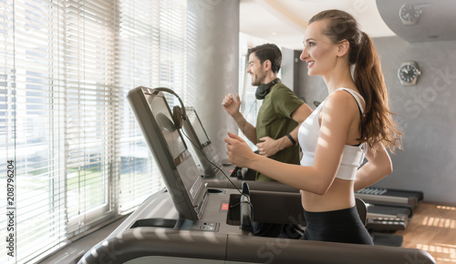 Happy young woman listening to music while running beside a handsome man on a modern treadmill with touch screen and headphones