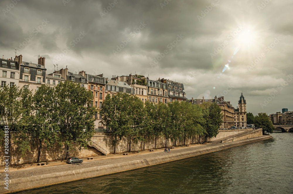 Old buildings, sidewalk and lined trees on the Seine River with sunlight in Paris. Known as the “City of Light”, is one of the most impressive world’s cultural center. Northern France. Retouched photo