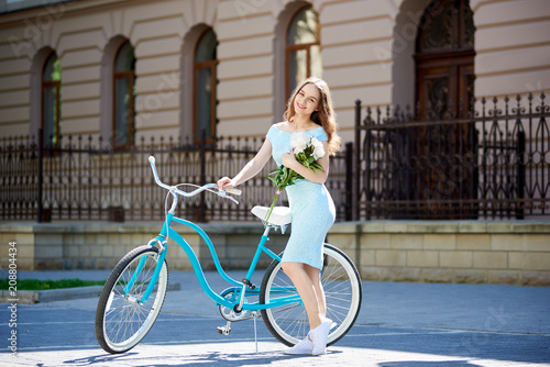 Smiling attractive female carrying flowers and posing next to blue bicycle in front of beautiful historical building during summer day. Pretty woman flowers turquoise bike fence house red doors