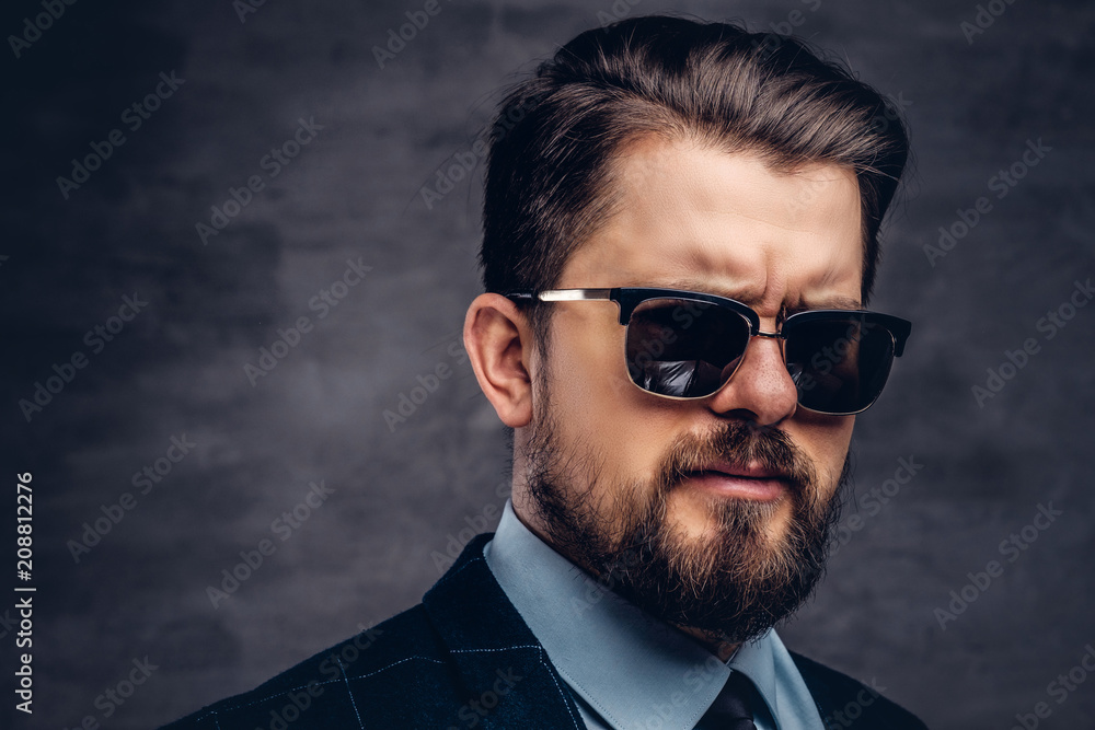 Close-up portrait of a handsome fashionable middle-aged man with beard and hairstyle dressed in an elegant formal suit and sunglasses. Isolated on a textured dark background in studio. Close-up portrait of a handsome fashionable middle-aged man with beard and hairstyle dressed in an elegant formal suit and sunglasses. Isolated on a textured dark background in studio.
