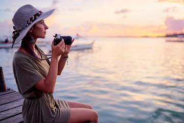 Photography and travel. Young woman in hat holding camera sitting on wooden fishing pier with beautiful tropical sea view.