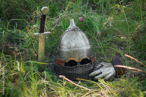  A helmet, a sword and a shield of a Mongolian warrior lie on the ground in a tall green grass.  Lost, taken after the battle. Medieval weapons of a nomad, steppe soldier.