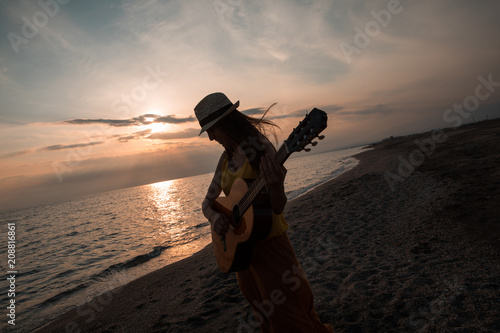 Silhouette of a young woman playing the guitar and dancing in the beach during a nice sunset. Musician and singer concept.