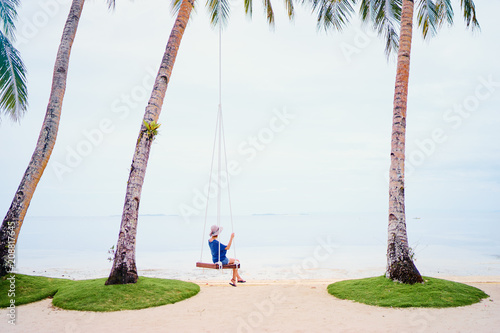 Vacation concept. Happy young woman in hat swinging at palm grove enjoying sea view. photo