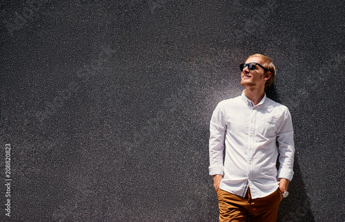 Handsome confident caucasian young man in sunglasses and white shirt leaning on grey wall.