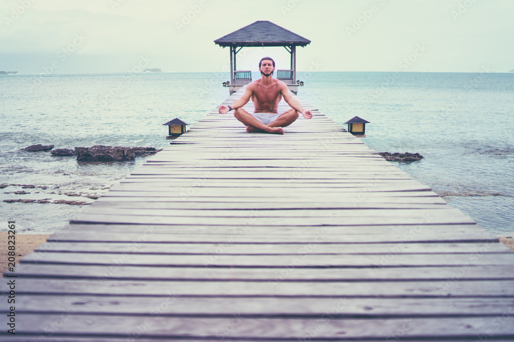 Yoga near the sea. Young man in lotus pose sitting on wooden pier. Concept of pray and meditation.