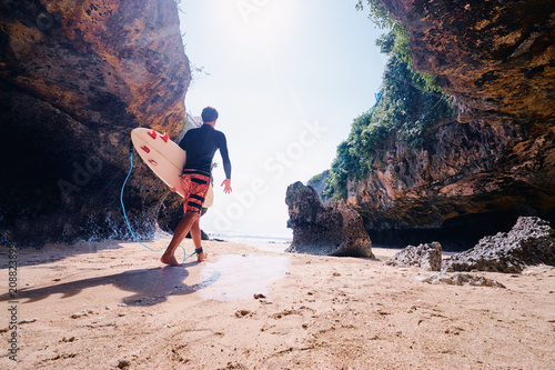 Hobby and vacation. Young man with surfboard on beautiful beach with high rocks. Uluwatu spot, Bali island, Indonesia.