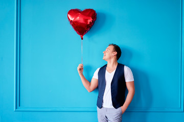 Saint Valentine's Day concept. Handsome young man holding heart shaped baloon against blue wall.