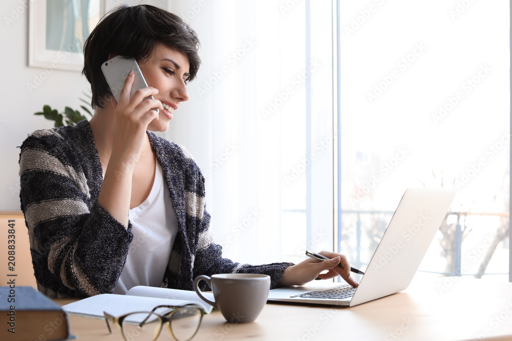 Obraz premium Young woman talking on mobile phone while working with laptop at desk. Home office