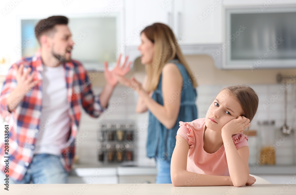 Little unhappy girl sitting at table while parents arguing on kitchen Little unhappy girl sitting at table while parents arguing on kitchen
