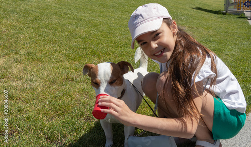 A girl sitting on a green grass in a cap gives the dog to drink water. Jack Rassell.