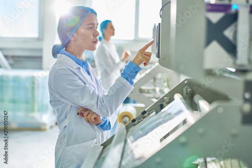 Side view portrait of young female worker wearing lab coat and hair cover standing by  power units pressing buttons on control panel in clean production workshop.