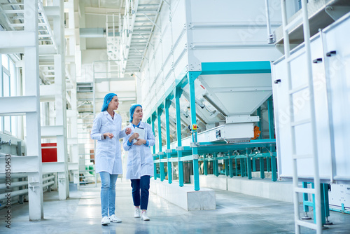 Full length portrait of  two young female workers wearing lab coats walking by  power units crossing hall in  in clean production workshop, copy space