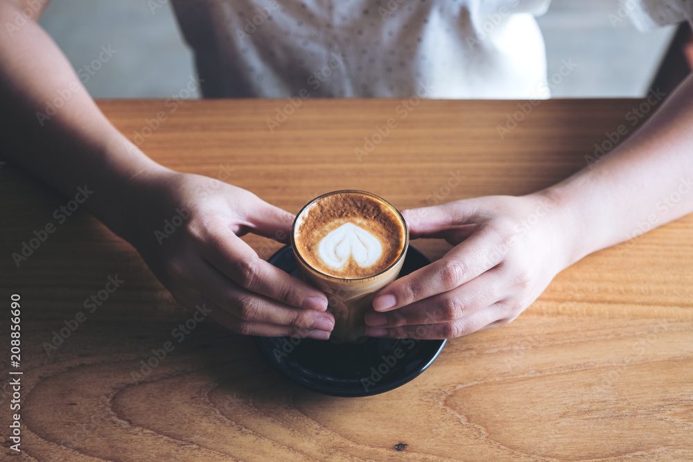 Closeup image of woman's hands holding a glass of hot coffee with heart latte art on wooden vintage table